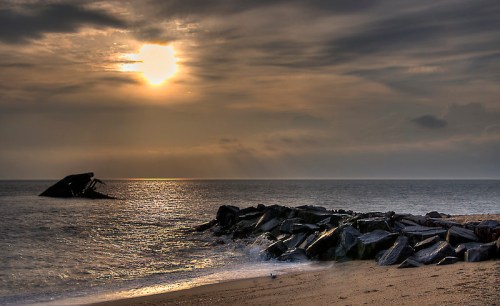 A serene beach scene at sunset, with rocky formations and a shipwreck silhouetted against a cloudy sky.