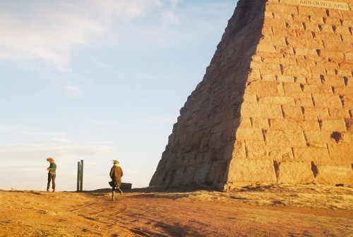 Two people stand near a large stone pyramid under a blue sky, with a rugged landscape in the background.