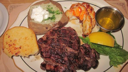 A plate featuring grilled steak, lobster tail, baked potato with sour cream, garlic bread, and a lemon wedge.