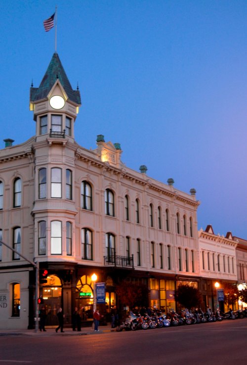 Historic building with a clock tower, illuminated at dusk, surrounded by parked motorcycles and streetlights.