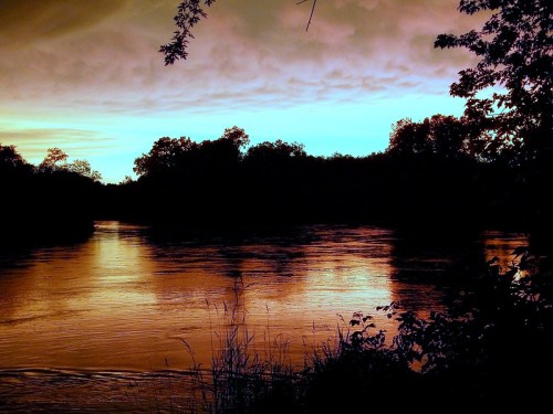 A serene river at sunset, reflecting vibrant orange and blue hues, framed by dark silhouettes of trees.
