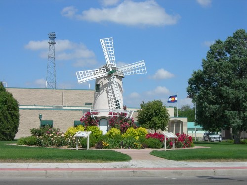 A windmill surrounded by colorful flowers and greenery, with a building and Colorado flag in the background under a blue sky.
