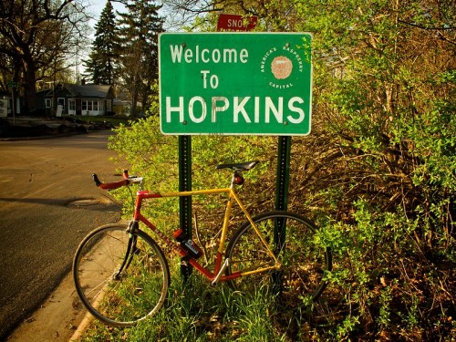 A yellow and red bicycle leans against a green "Welcome to Hopkins" sign surrounded by greenery.