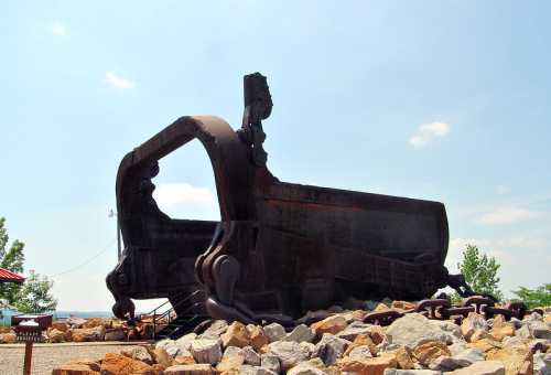 A large, rusted mining bucket sits on a rocky base, surrounded by greenery and under a clear blue sky.