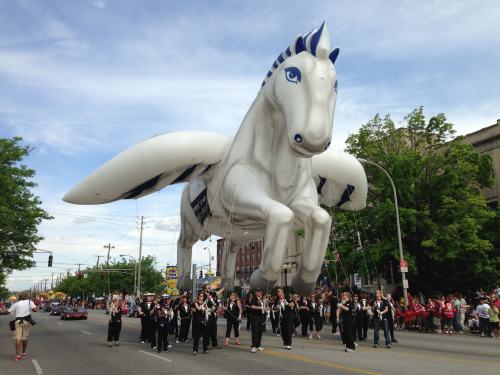 A large white horse balloon floats above a marching band in a parade, with trees and spectators in the background.