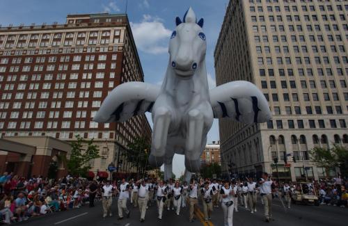 A large inflatable horse floats above a parade, with people in white shirts marching below in a city street.