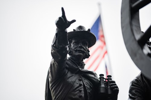 A close-up of a bronze statue of a man in a hat, holding binoculars, with an American flag in the background.