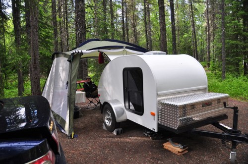 A small white camper trailer set up in a forested campsite, with an awning and outdoor seating area.