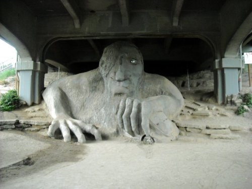 A large stone troll sculpture sits under a bridge, with a rugged face and one hand holding a small object.