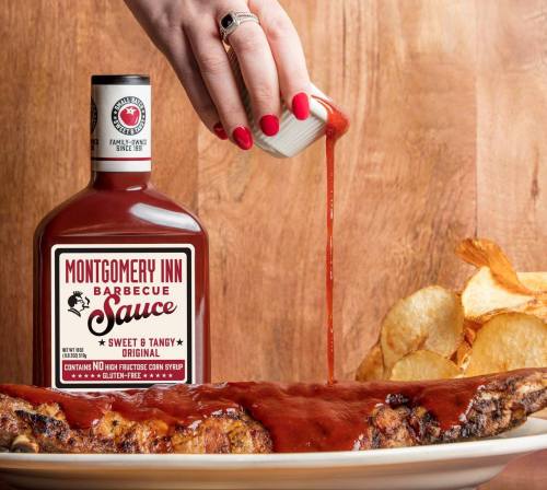 A hand pours Montgomery Inn barbecue sauce over ribs, with chips and a wooden background.