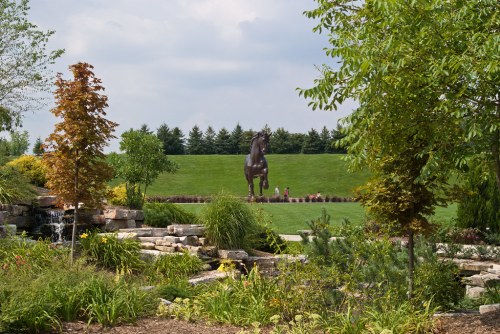 A lush garden with a waterfall and colorful plants, featuring a large horse statue in the background against a green hill.