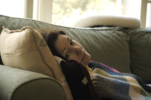 A person resting on a couch, surrounded by pillows and a colorful blanket, looking contemplative.