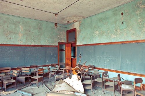 Abandoned classroom with dusty, overturned chairs and peeling blue walls, featuring chalkboards covered in writing.