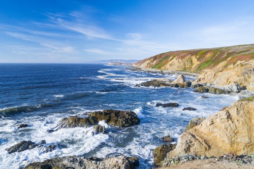 A scenic coastal view with rocky cliffs, waves crashing against the shore, and a clear blue sky.
