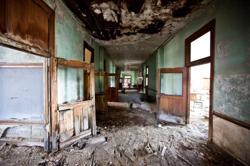 Abandoned hallway with peeling green walls, broken windows, and debris scattered on the floor.