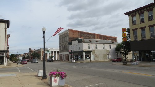 A small town street scene featuring historic buildings, an American flag, and flower planters along the sidewalk.