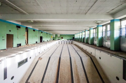 Abandoned indoor swimming pool with peeling green walls and a wooden floor, illuminated by natural light.