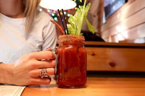 A hand holding a large glass mug of a cocktail garnished with celery and bacon, set on a wooden table.