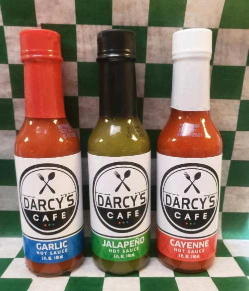 Three bottles of Darcy's Cafe hot sauce: Garlic (red), Jalapeño (green), and Cayenne (orange) on a checkered background.