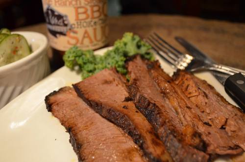 Sliced brisket on a plate with kale and pickles, accompanied by a bottle of barbecue sauce in the background.