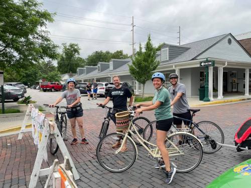Four people on bicycles, wearing helmets, smile while standing on a brick road near shops and trees on a cloudy day.