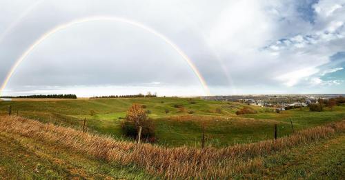 A panoramic view of rolling green hills under a double rainbow and a cloudy sky.