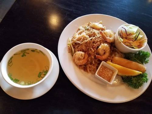 A plate of shrimp pad thai with salad, spring rolls, and a bowl of clear broth on a dark table.