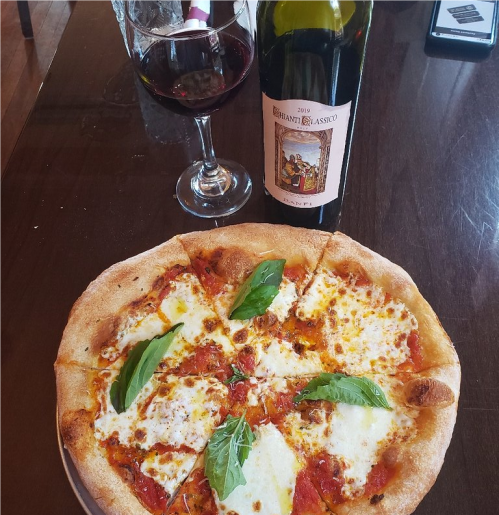 A freshly baked pizza topped with mozzarella and basil, accompanied by a glass of red wine and a bottle on the table.