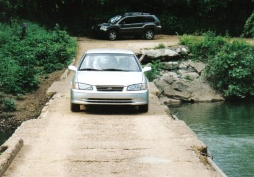 A silver car drives cautiously on a narrow, rocky bridge over a river, with greenery surrounding the area.