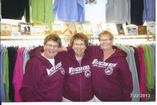 Three women wearing matching maroon "Wisconsin" hoodies smile together in a clothing store, surrounded by colorful apparel.