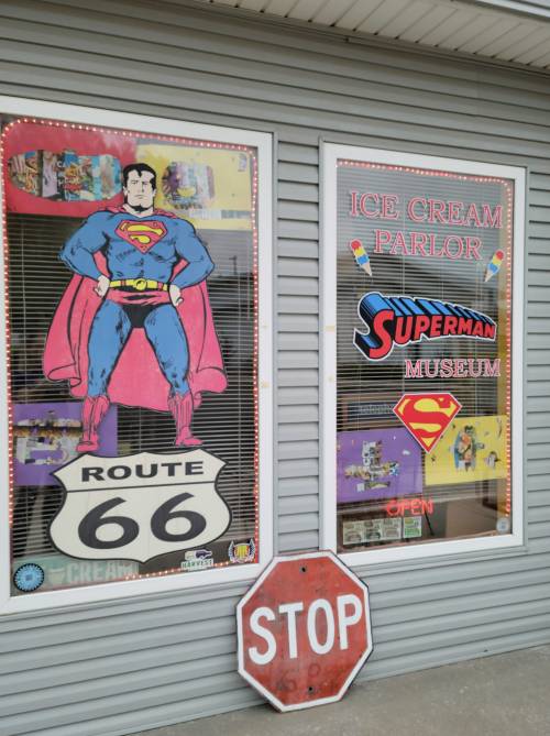 A storefront featuring Superman artwork, an ice cream parlor sign, and a Route 66 sign, with a stop sign in front.