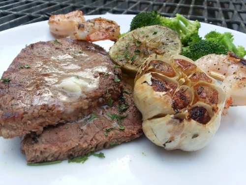 A plate featuring grilled steak, shrimp, roasted garlic, and steamed broccoli, garnished with herbs.
