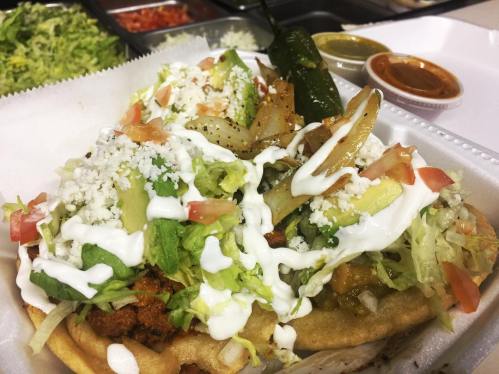 Two loaded tacos topped with lettuce, tomatoes, cheese, sour cream, and grilled onions, served with sauces on the side.