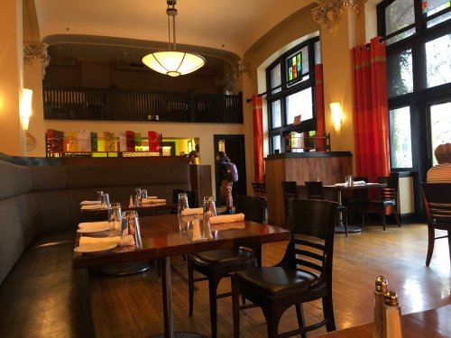 Interior of a restaurant featuring empty tables, large windows, and colorful decor, with a person walking in the background.