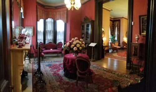 A vintage parlor with red walls, ornate furniture, and a floral centerpiece on a round table, featuring rich rugs and curtains.