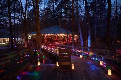 A wooden walkway adorned with colorful lights leads to a cozy, illuminated pavilion surrounded by trees at dusk.