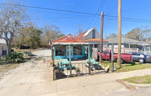 A small, colorful building with outdoor seating, surrounded by trees and parked cars on a sunny day.