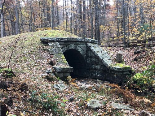 A stone arch bridge over a small stream, surrounded by autumn foliage and a forested landscape.