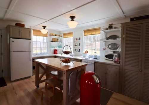 Bright kitchen with a wooden island, white cabinets, and large windows, featuring a red retro fridge and organized shelves.