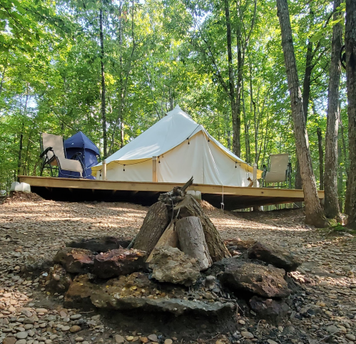 A glamping tent on a wooden platform surrounded by trees, with a small campfire in the foreground.