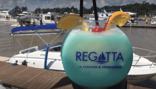 A colorful cocktail in a large glass with fruit garnishes, set against a marina with boats in the background.
