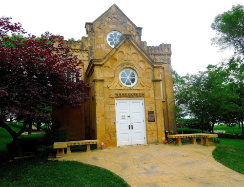 Historic yellow stone building with ornate architecture, surrounded by green trees and a paved walkway.