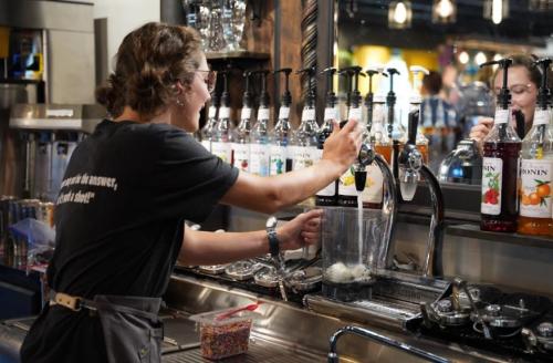 A barista prepares a drink at a coffee shop, using syrup dispensers and a blender in a busy café setting.