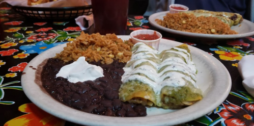 A plate of enchiladas topped with green sauce and sour cream, served with rice, black beans, and salsa on a floral tablecloth.