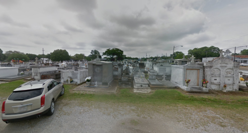 A cemetery with numerous gravestones and mausoleums under a cloudy sky, with a parked car in the foreground.