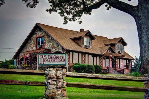 A stone and wood house with a sign reading "R.S. Biggers Homestead, Established 1918," surrounded by greenery.