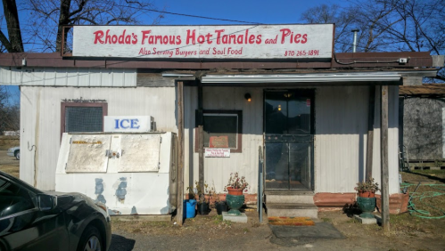 A small, rustic building with a sign reading "Rhoda's Famous Hot Tamales and Pies," offering burgers and soul food.