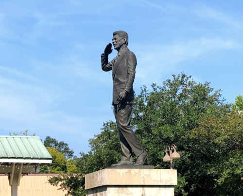 A bronze statue of a man in a suit saluting, standing on a pedestal surrounded by trees and a clear blue sky.