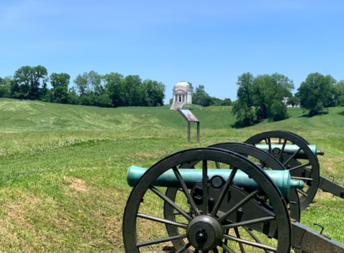 Two cannons in the foreground with a grassy landscape and a monument in the distance under a clear blue sky.