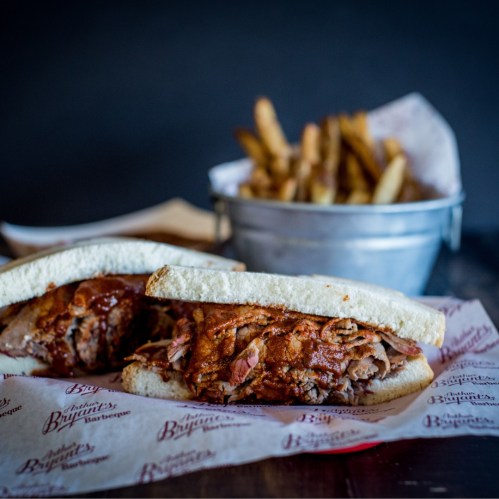 A close-up of a barbecue sandwich with pulled meat on white bread, served with a side of fries in a metal bucket.